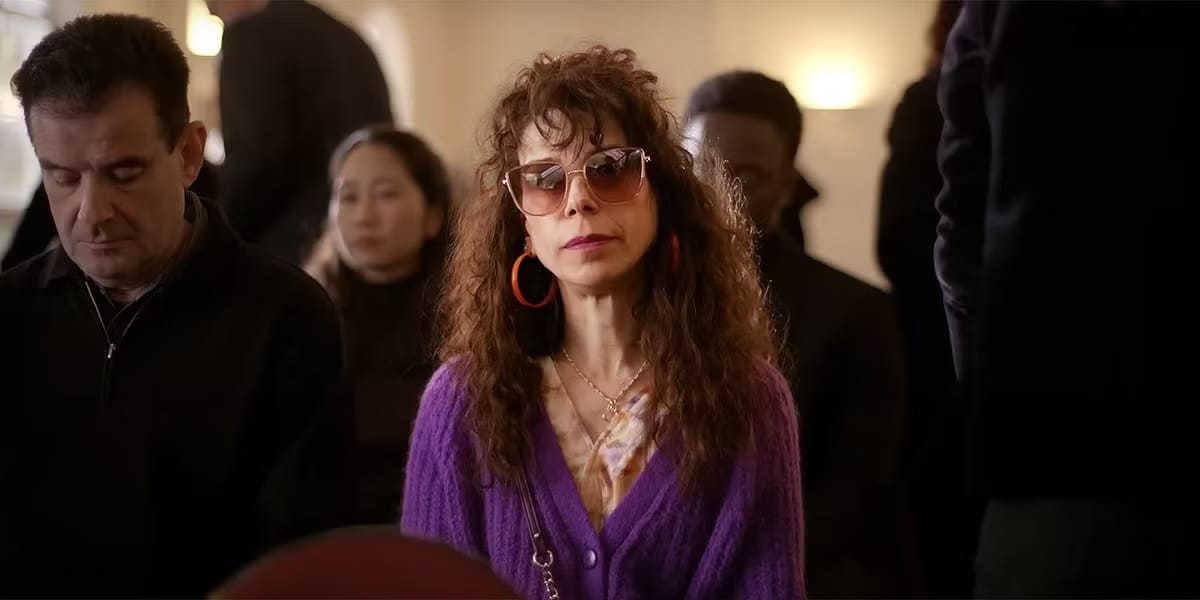 Sally Hawkins in a still from Bring Her Back, wearing a bright purple sweater with huge frizzy hair, gigantic hoop earrings, and rock star sunglasses, surrounded by a bunch of sad people all in black. Instantly iconic